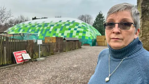 Jackie Christie Green stands near the huge inflatable. He has gray short hair and glasses. He is wearing a blue jumper with a silver pendant necklace at the top.