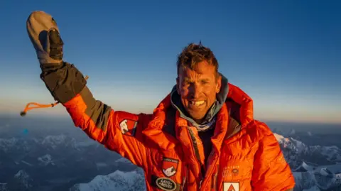 Kenton Cool A man wearing a bright orange puffer jacket with badges on it raises his gloved hand in the air as he stands at the summit of Everest. The snow-capped peaks of other mountains can be seen behind him and the light is golden, as if it is sunrise.