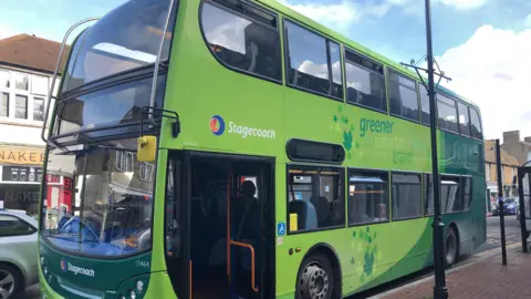 A green bus at a bus stop. It is branded with the company name - Stagecoach. It is parked near a bus stop and there are shops in the background