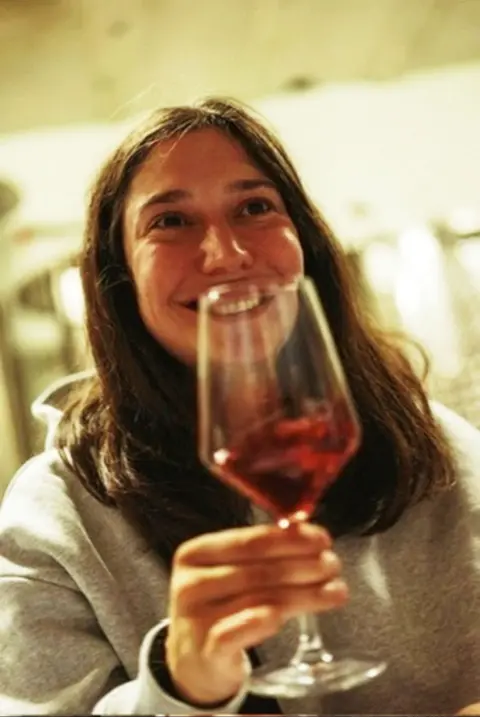 Holly Willcocks Glass containing red wine held in the foreground by a person seated indoors. The person is wearing a light-coloured sweatshirt. The glass is positioned close to the camera, with the person and a softly lit interior visible in the background.
