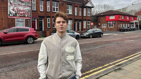 Man in beige jacket stands on a roadside. He has ginger hair and is in his early 20s.