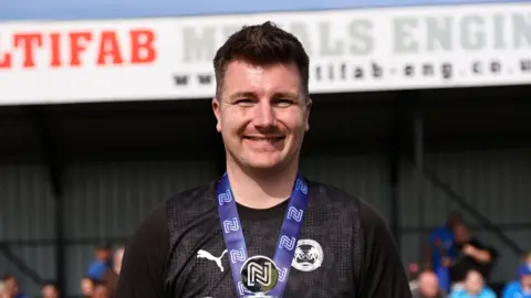 DARREN WILES Jake Poole is smiling and looking at the camera. He is wearing a black football top and medal. Behind him is a football stand of fans, blurred. 