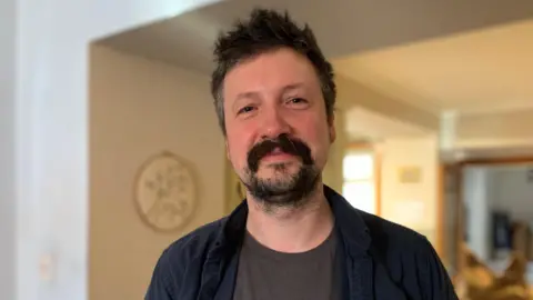 A man with dark spikey hair and a beard in a blue shirt with a grey T-shirt underneath