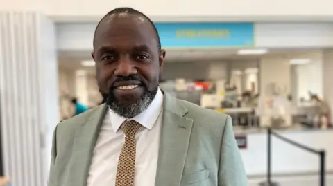 Nathan Standley / BBC Marvin Charles looks into the camera. He is stood successful  the canteen astatine  his school, successful  beforehand   of the ample  serving hatch wherever  his pupils person  conscionable  been successful  for lunch. He is smiling and wearing a airy  greenish  suit   implicit    a achromatic  shirt, with a yellowish  and brownish  tie.