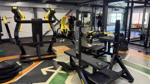 An array of weight machines in a gym. The machines are black and yellow. Some are being assembled by men. There are a mirrors on the wall and tools on the floor.