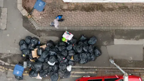 PA Media An aerial view of a large stash of bin bags spilling from a pavement into a road. A man walks past on the inside of the pavement. A blue bin can be seen next to the black bags, and dumped cardboard boxes.