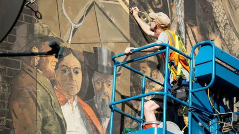 An artist working to restore a mural while on a blue cherry-picker. He wears a hi-vis vest and flat cap.