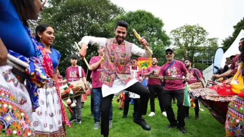 PA Indian dancers and musicians in traditional clothing are performing in a park. they're standing on grass.