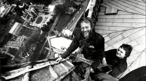 Dawson Steeplejacks A black and white photo of two men looking up at a camera as they are harnessed to a ladder on the pinnacle of a cathedral. The city is in the distance beneath them.