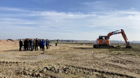 Luke Deal/BBC A group of people stand in a large field close to where a digger rests. 