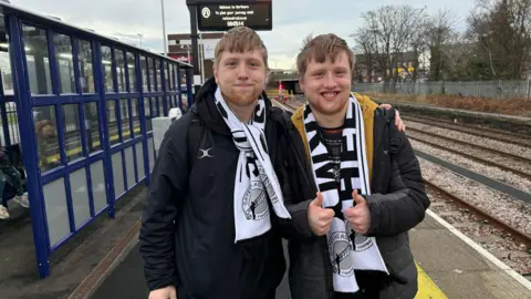 Twin brothers James and Thomas Lovejoy are wearing black and white Gateshead football club scarfs. They have light brown hair and smiling to the camera. They are standing on a station platform. 