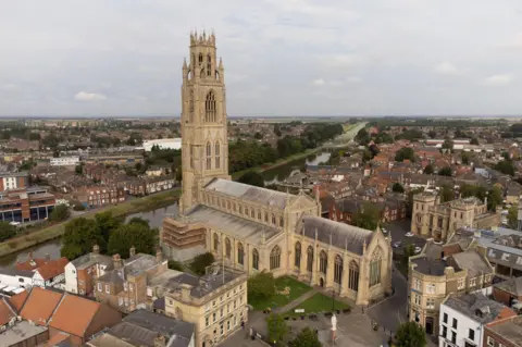 A drone view of the centre of Boston, including a large 16th Century gothic church that dominates the skyline with a tall, ornate tower. Other grand buildings surround the church and there are red-brick houses and shops to either side. A river runs to the left of the church. The sky is grey.