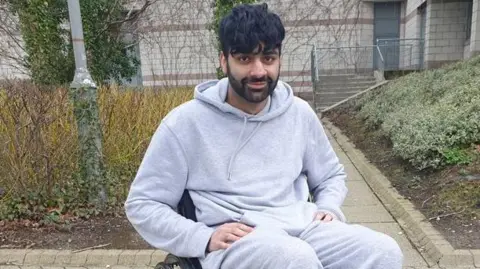 A young man with dark hair and facial hair sits, smiling at the camera. He is dressed in a grey hoodie and sweatpants. He is outside.