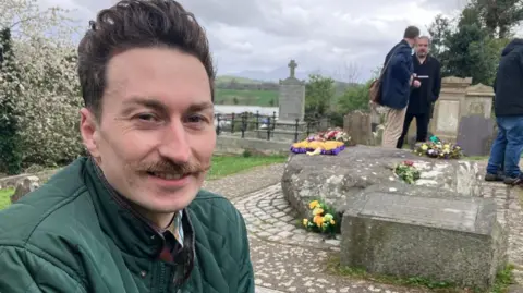 A man with brown hair and a moustache sits in the foreground of a graveyard beside a stone grave adorned with flowers, he wears a green jacket. Other people stand and talk nearby among gravestones, with rolling countryside visible in the background under a cloudy sky. 