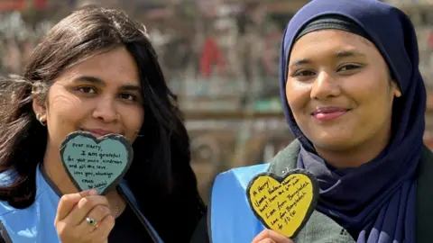 Holmes Chapel Partnership Two young women smile at the camera holding slate hearts with messages to Harry Styles