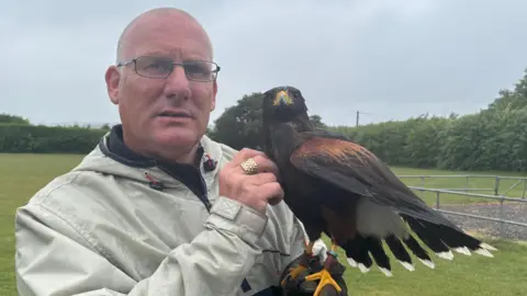 Wayne Housden is standing outside holding the Harris hawk. He is wearing a beige coat and is looking directly at the camera and stroking the bird.
