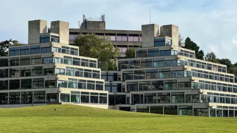 The pyramid-shaped Ziggurats at the University of East Anglia can be seen, with their angular concreate and glass structures in view, with grass in the foreground.