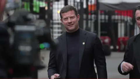 A man with short brown hair wearing an all black suit with a black polo neck and a little shamrock pin on his lapel walks towards a waiting photographer