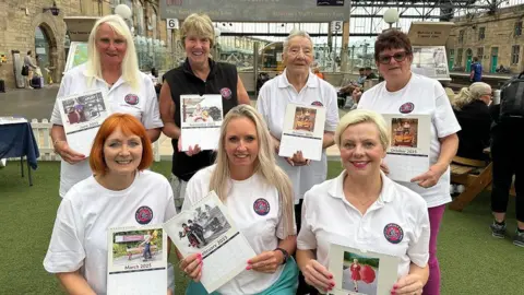 Settle-Carlisle Railway Development Company Seven women of different ages standing at the train station with their calendars. A sign in the background reads Welcome to Settle-Carlisle Line.