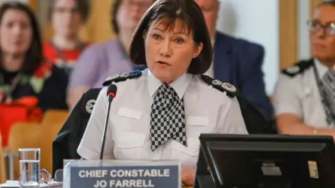 Jo Farrell, who has short brown hair, wearing a white police uniform with black badges sewn on the shoulders speaks into a microphone as people site behind her in the public gallery. There is a sign on the desk in front of her with her name and rank