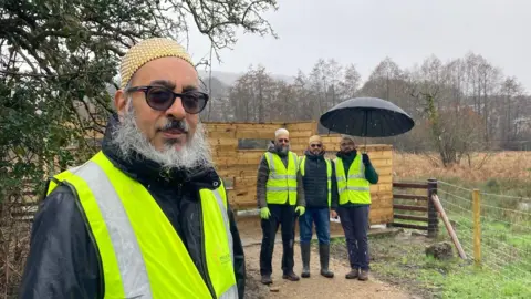Four men in hi-vis jackets with one holding a black umbrella standing next to a wooden bird observation deck 