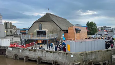 Andrew Turner/BBC An old brick building with a thatched roof on Great Yarmouth's waterfront. A modern metal balcony area has been build on one end, with a wooden extension on one side. There is a shipping container next to the harbour wall and lots of people milling around.