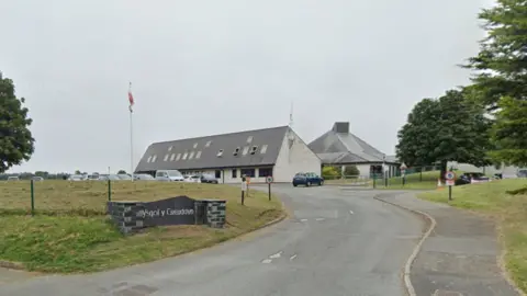 Google A screenshot from Google Maps showing the primary school, Ysgol y Creuddyn. Looking at the white building from the point of view of the main road. A Welsh flag hangs on a pole in the distance. Trees either side of the school.