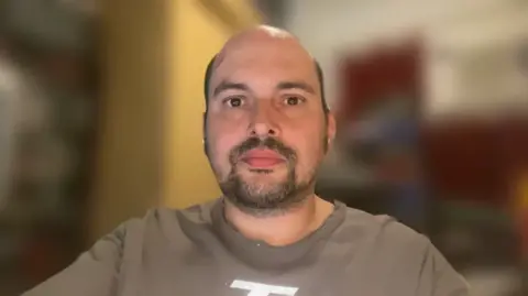 BBC Frank Schuengel in a brown t-shirt. He has dark stubble and dark eyes. The background is blurred.