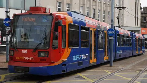 A Sheffield Supertram moves through the city's streets. It is red orange and blue and has the word "Halfway" on its information board. 