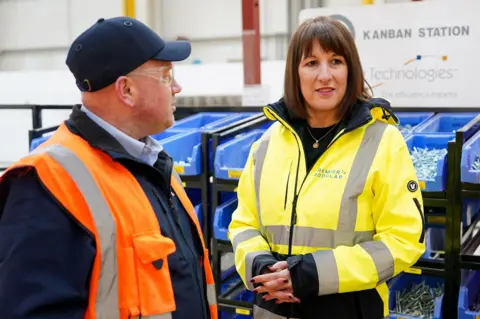 Rachel Reeves (kanan), mengayunkan mantel tahan air kuning, berdiri di samping seorang pria dengan topi dan jaket hi-vis di sebuah acara pada hari Kamis.