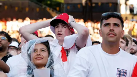 PA Media England fans watch a screening of the World Cup 2026 semi-final match between England and Argentina. A woman (left) with brown hair wearing a plastic see-through rain cover holds a drink, a man (centre) behind her has his hands on his head in despair and a man on the right looks intently at the screen.