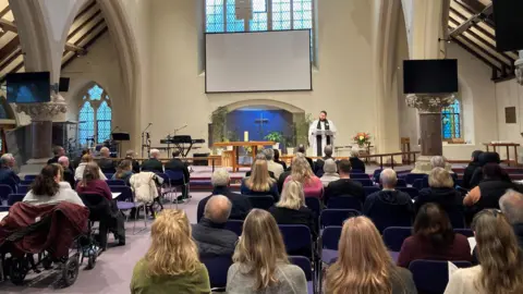 A small congregation sitting in a church with their backs to the camera, with a vicar speaking at the front.