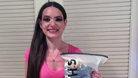 Deborah Grushkin, her eyes dramatically outlined, wears a pink shirt and holds up a Shein package