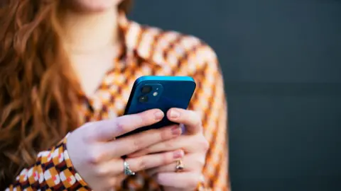 Getty Images The hands of a woman holding a phone. She can be seen in the background wearing a black and orange top. Her face is out of shot, but she has long red hair. 