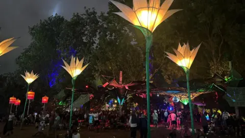 About six flower-themed lanterns can be seen dotted around an area of Glastonbury Festival. They are about the height of a street light, with green "stems" and yellow flowers. There is an under-tree canopy in the background with a disco ball hanging for it, with revellers dancing underneath it.
