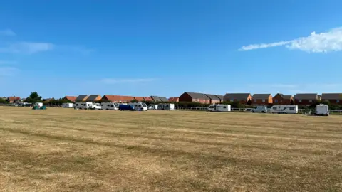 More than 10 white caravans have camped on a field of dry grass in front of a housing estate in Redcar. The caravans are spread out along the far side of the field and the sky is clear and blue.