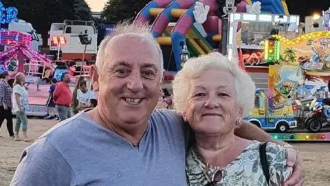 Bill Sheargold A man and a woman standing arm in arm in front of a fairground