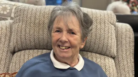 Betty Redfern smiles at the camera while sitting in a beige armchair in her care home. She has short, grey hair and is wearing a blue sweatshirt over a white top with a soft collar. 