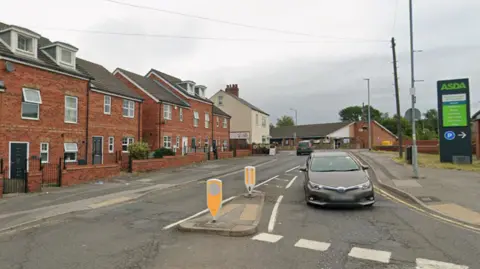 A Google streetview image of St. Luke's Road, near the junction with High Street and Queensway, in Grimethorpe, Barnsley. A car drives towards the camera. On the left hand side of the road is a row of modern brick-built houses. On the right is a sign for a nearby Asda supermarket.