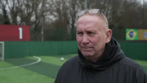 BBC Alana Knight has short white hair, and has glasses sat on top of his head. He is wearing a black coat and is standing on a caged artificial football pitch.