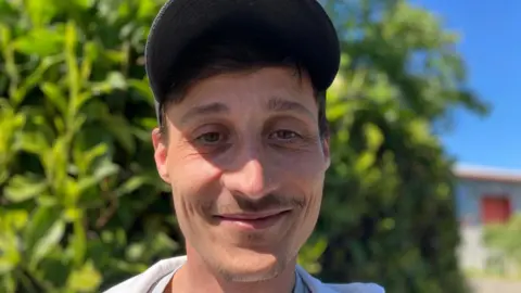 A head and shoulders photograph of Andrew Nicoll who is wearing a grey t-shirt, white hooded top and a black baseball cap.