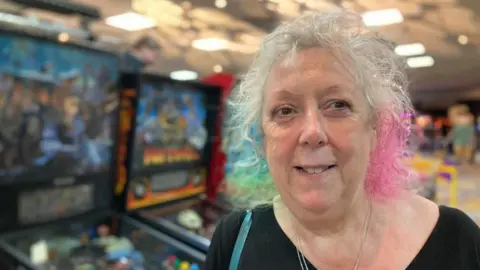 Martin Heath/BBC Ailsa Cunie with green, blue and pink hair wearing a black top and standing in front of pinball machines
