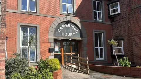 There is a ramp leading to the entrance to Cambridge Court Care Home. There are plants and bushes at the front of the red-bricked building. 