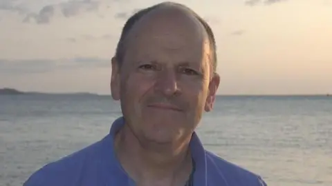 University of East Anglia Robert Nicholls smiles at the camera. Behind him, the sea can be seen during sunset. He has short, shaved hair and wears a blue polo shirt. 