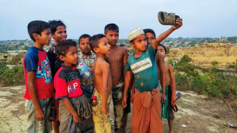 Children pretending to take a selfie at a refugee camp by using a sandal as a phone