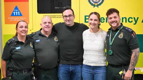 London Ambulance Service Paramedics stand next to Patrick and Tash during the reunion. They are standing by an ambulance.