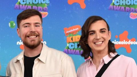 MrBeast pictured with Ava Kris Tyson at the Nickelodeon Kids Choice Awards. MrBeast has short brown hair and a short beard and wears a white overshirt over black T-shirt. Ava has shoulder-length brown hair parted to the side and wears a pink shirt.