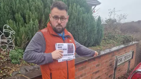 Local Democracy Reporting Service A man, wearing glasses, in an orange top with dark hair and a beard is by a wall and looking at the camera. Bushes are behind the wall.