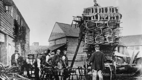 Wycombe Museum A black and white photo showing chairs being loaded onto a wagon by a group of men in caps, at Gibbons chair factory, off Oxford Road, High Wycombe in about 1895. 