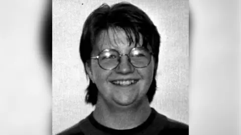 Hughes Family A black and white image of Julia Hughes who has short hair with a fringe, round metal glasses and is smiling at the camera. She is wearing a t-shirt.